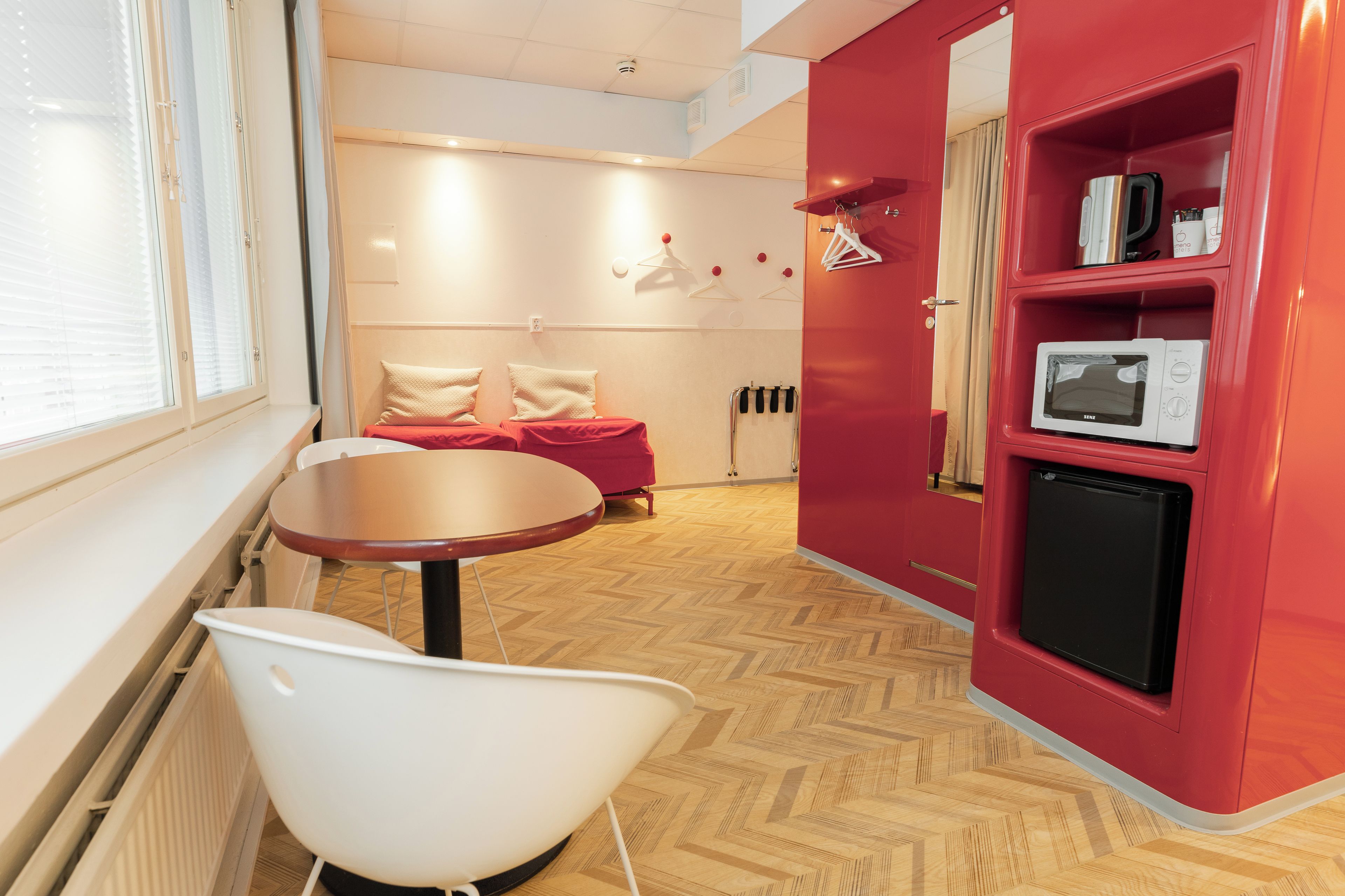A hotel room with a red wall, a white chair, a small table, a bed with red bedding, a microwave, and a heater.