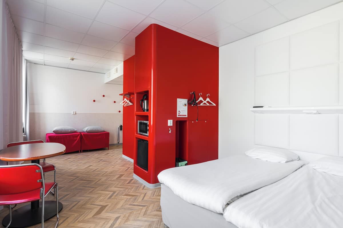 A modern hotel room with a red accent wall, a bed, a small kitchenette, a seating area with a table and chairs, and a view of the city.