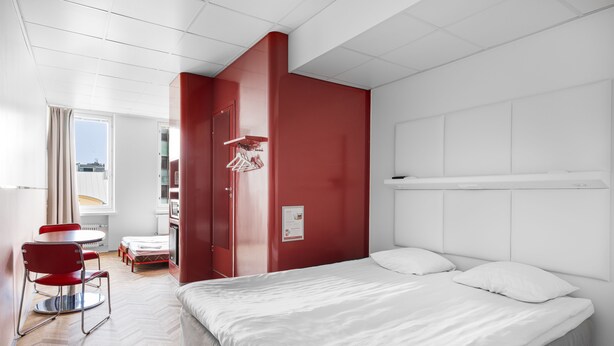 A modern hotel room with a large bed, a red accent wall, a dining area with a red chair, and a view of the city through the window. - Omena Hotel Tampere (Tampere)