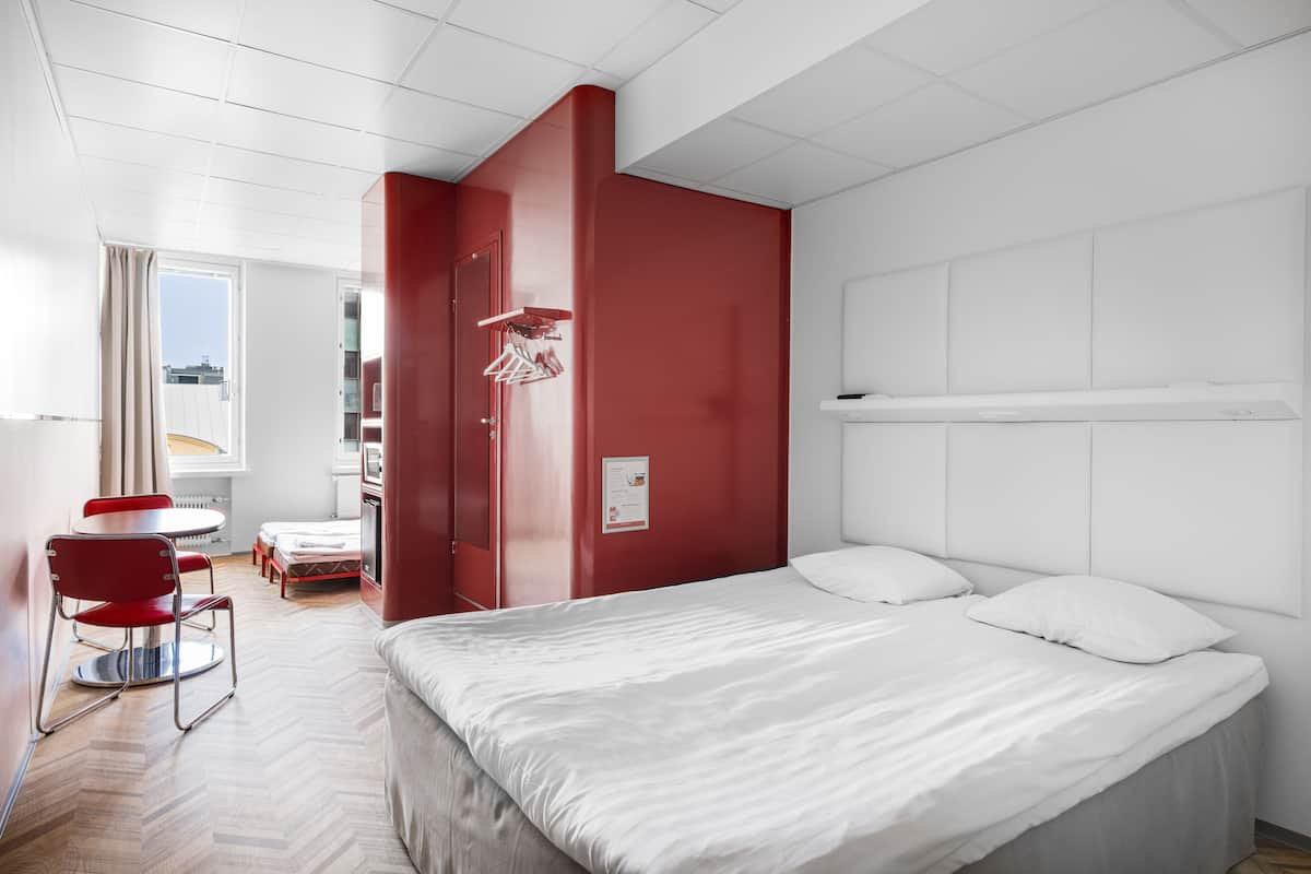 A modern hotel room with a large bed, a red accent wall, a dining area with a red chair, and a view of the city through the window.