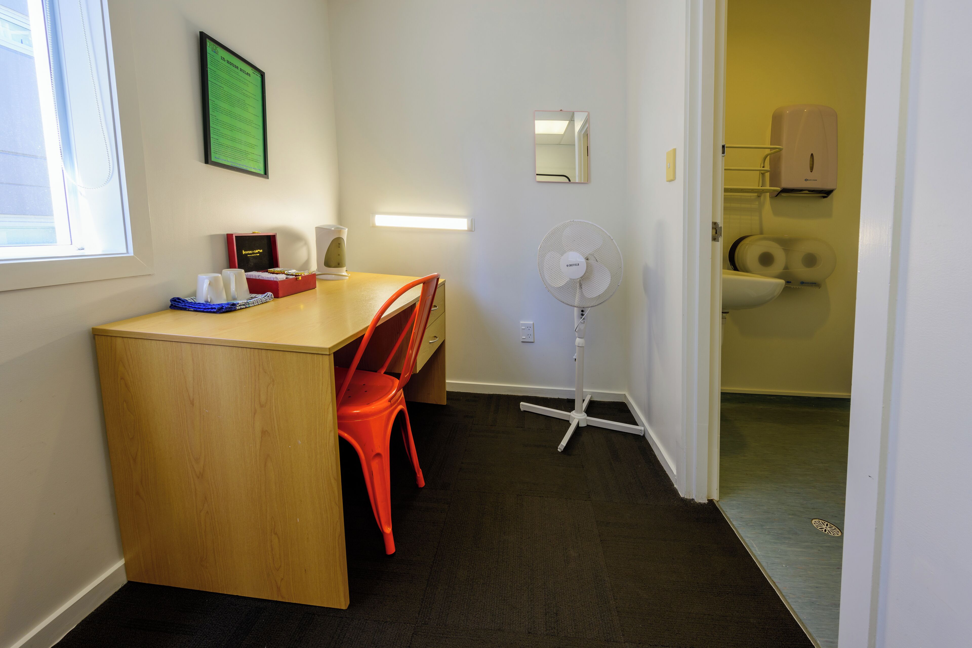 Hotellihuone, jossa on puinen työpöytä, punaiset tuolit, lattiatuuletin, wc ja käsidesin annostelija.