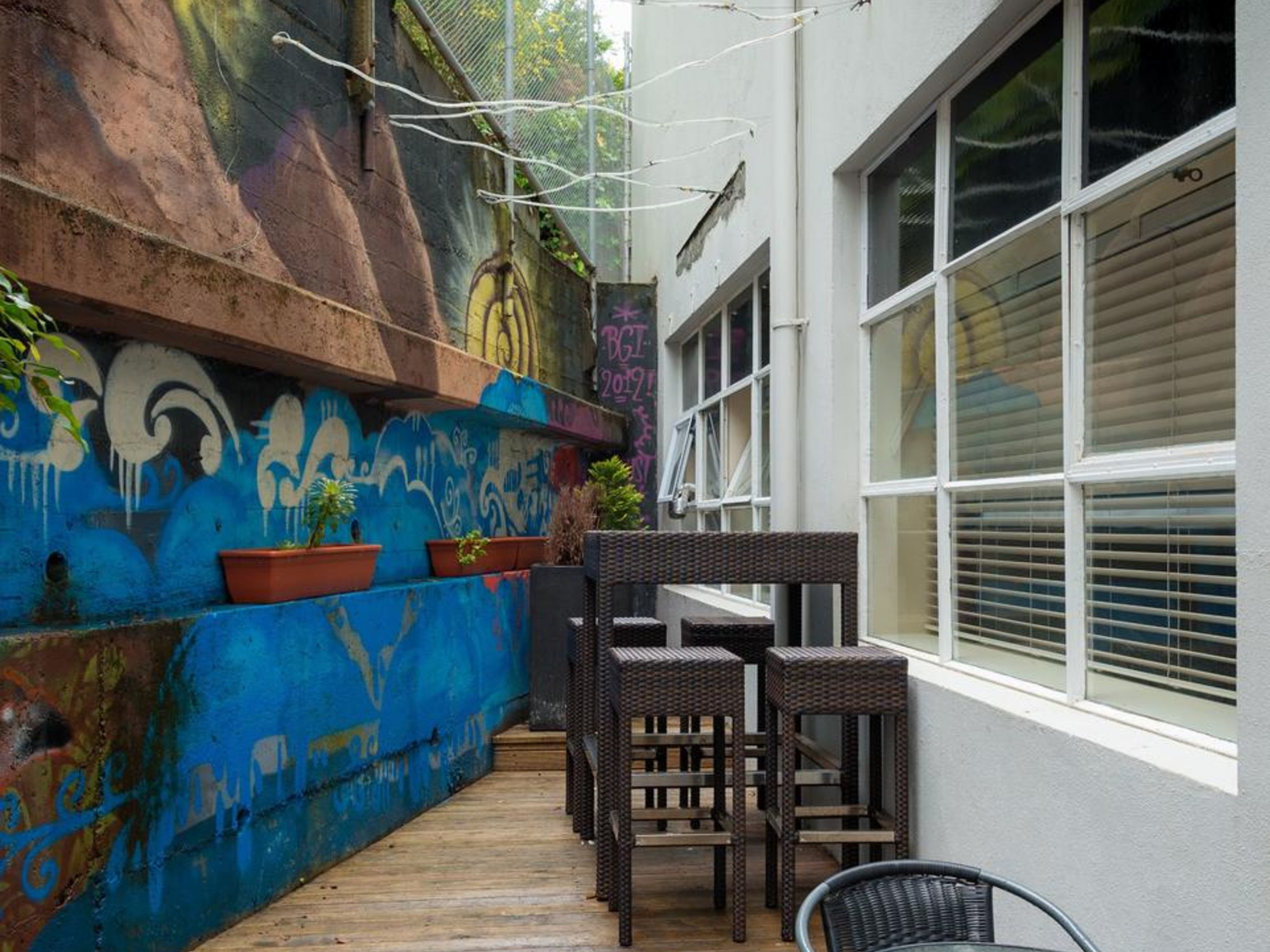 A narrow outdoor space with a wooden deck, wicker furniture, and a wall with colorful graffiti.