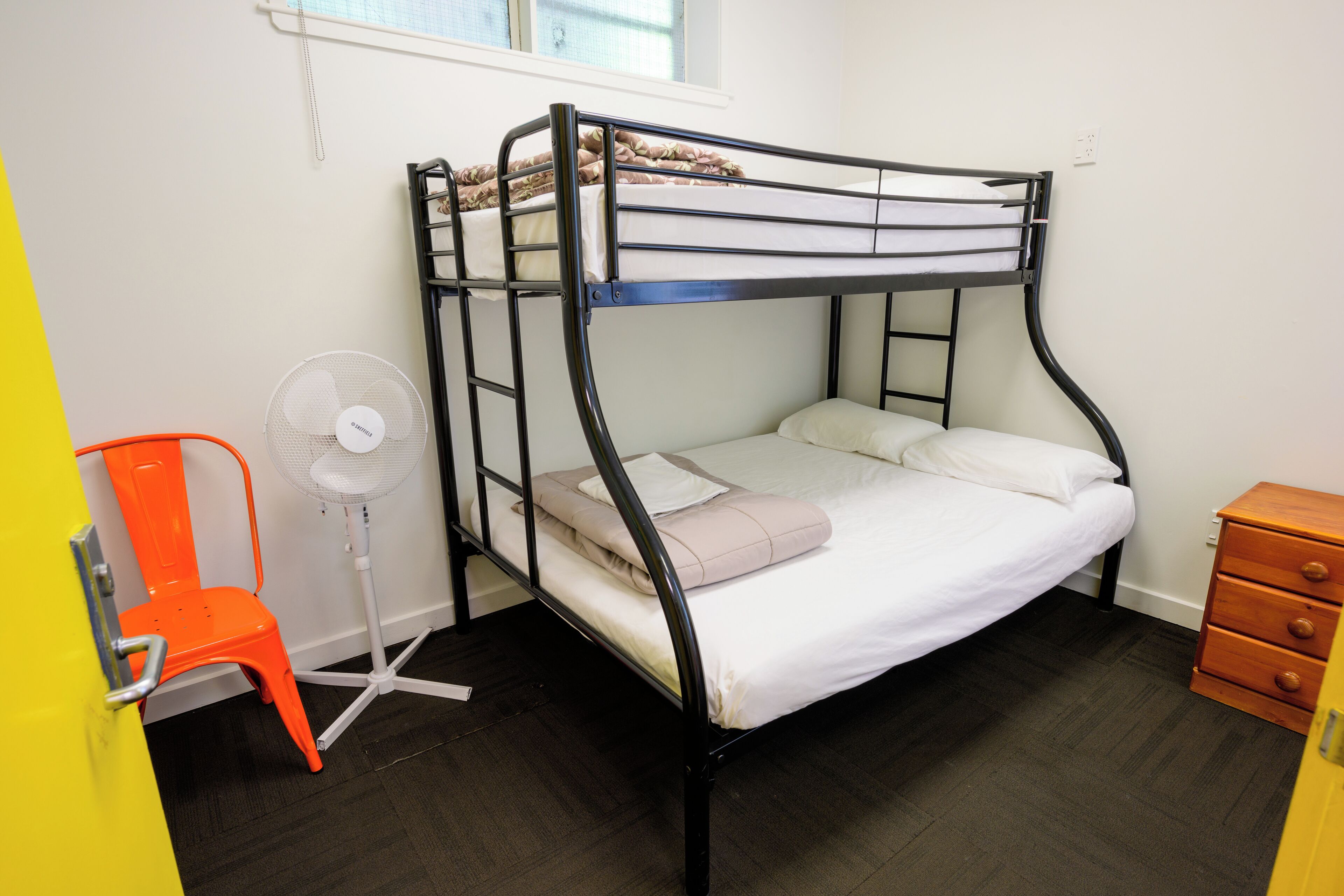 A bunk bed with a mattress and pillows, a small wooden dresser, a fan, and an orange chair in a room with a window and a door.