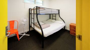 A bunk bed with a white mattress and a single pillow, a white desk chair, a white standing fan, and a wooden dresser.