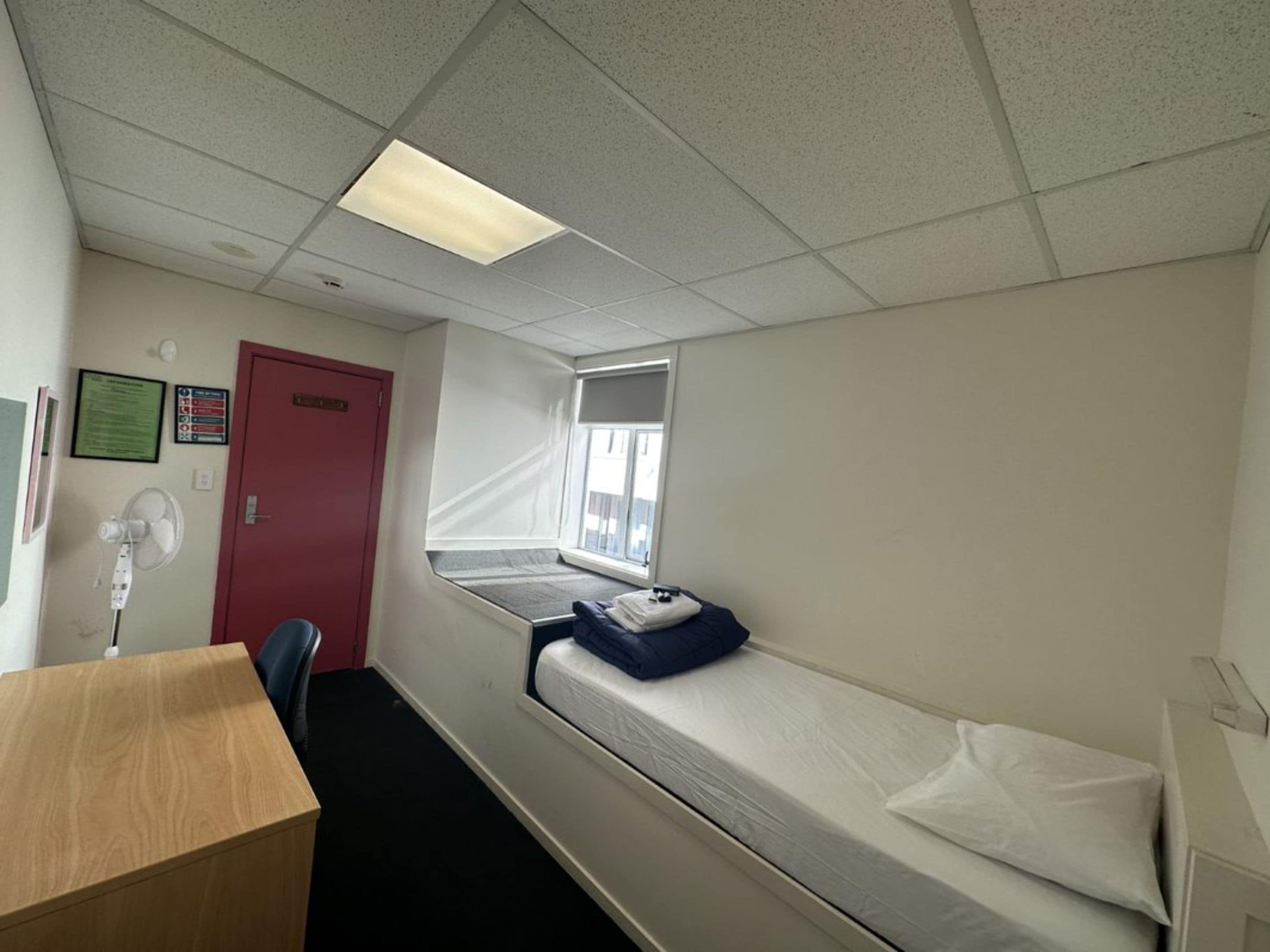 Ein Zimmer mit einem Einzelbett, einem Schreibtisch, einem Fenster mit Aussicht, einer roten Tür und einem Wandventilator.