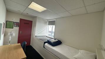 Ein Zimmer mit einem Einzelbett, einem Schreibtisch, einem Fenster mit Aussicht, einer roten Tür und einem Wandventilator.