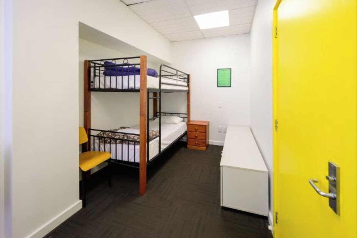 Ett studentrum med våningssängar, ett skrivbord, en stol, en liten byrå och en gul dörr.
