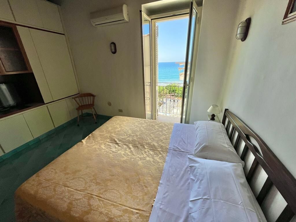 Quarto com cama, cadeira, guarda-roupa e vista para o mar através de uma porta.