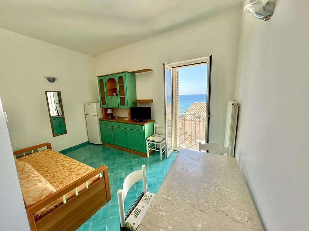 A compact room with a bed, a small kitchenette, a dining table, and a view of the sea through a sliding door.