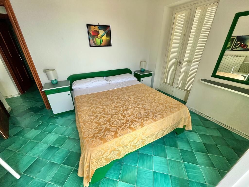 A room with a bed, a nightstand, a lamp, a painting, and a window with blinds.