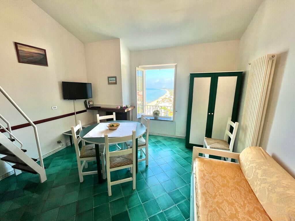 Una stanza piccola e luminosa con un tavolo da pranzo e sedie, una TV, una finestra vista mare e un termosifone.