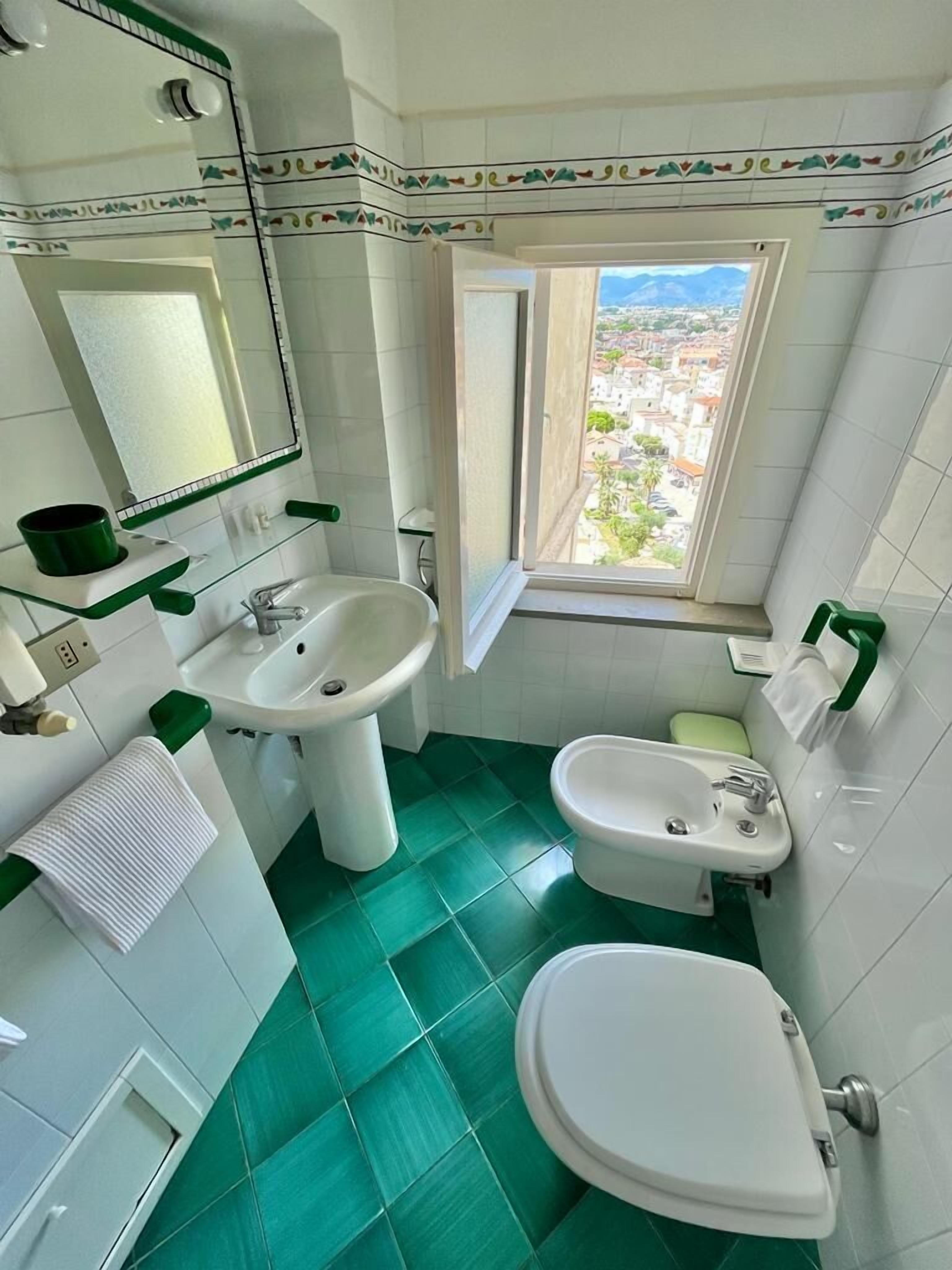 A bathroom with a toilet, bidet, and sink, green tiles, and a window with a city view.