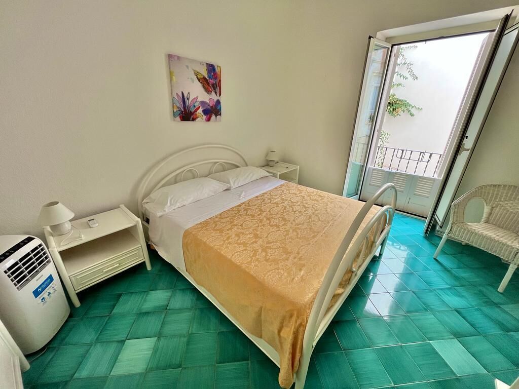 A bedroom with a bed, a nightstand, a lamp, a heater, a chair, and a window with a view of greenery.