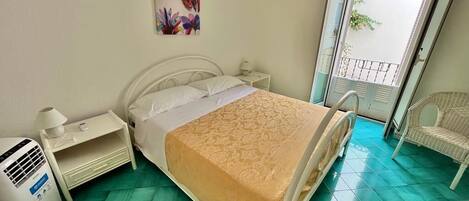 A bedroom with a bed, a nightstand, a lamp, a heater, a chair, and a window with a view of greenery.