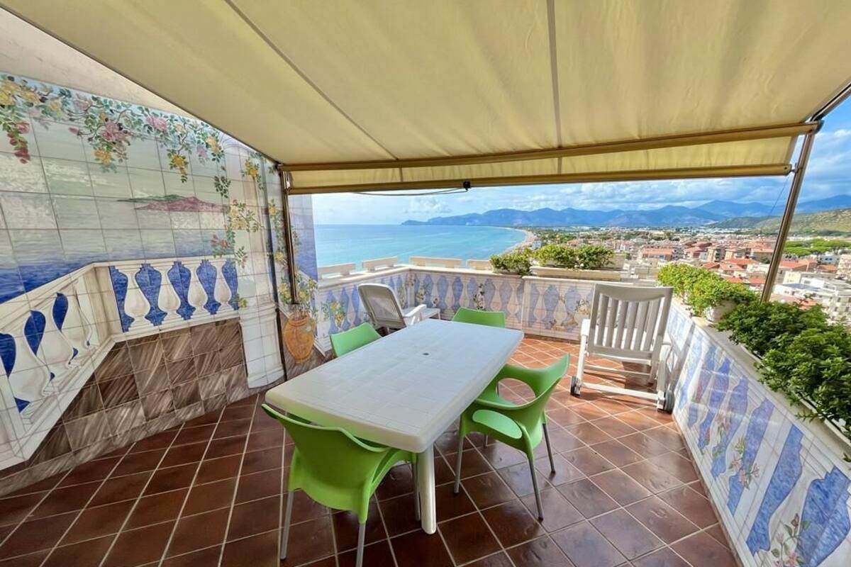 Eine überdachte Terrasse mit Tisch und Stühlen, eine Wand mit Fliesen und einem floralen Muster sowie Meer- und Landschaftsblick.