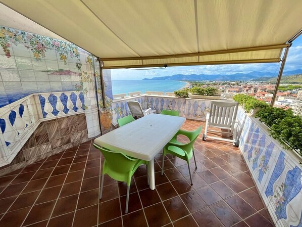 Una terraza techada con una mesa y sillas, un muro con baldosas y motivos florales, y vista al mar y el paisaje distante.