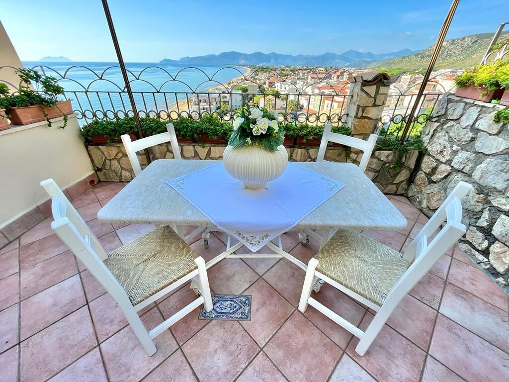 A patio with a table and chairs, a vase with flowers, and a view of the sea and mountains.