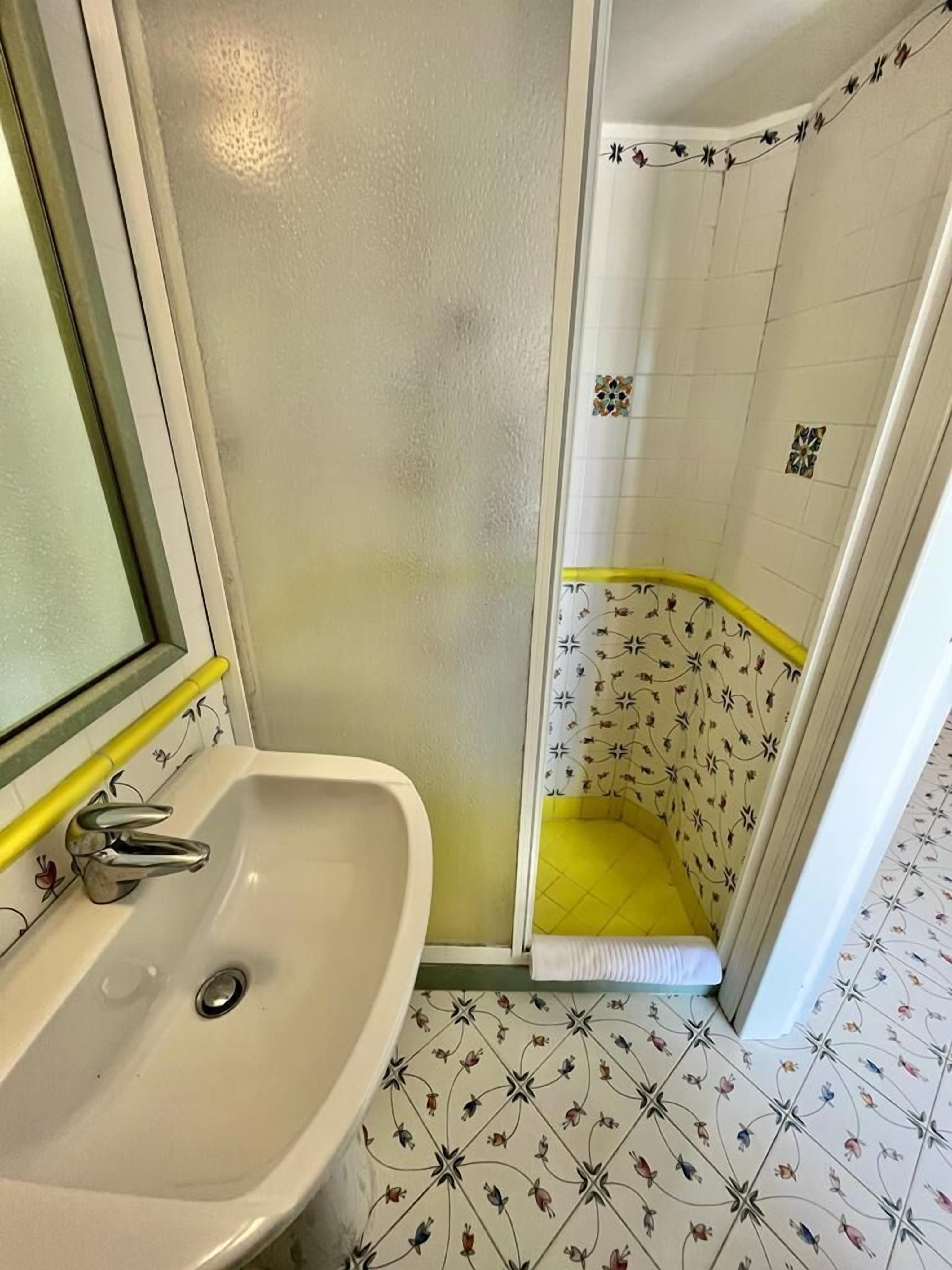 Ein Badezimmer mit einem weißen Waschbecken, einer Dusche mit floralen Fliesen und einer gelben Badmatte.