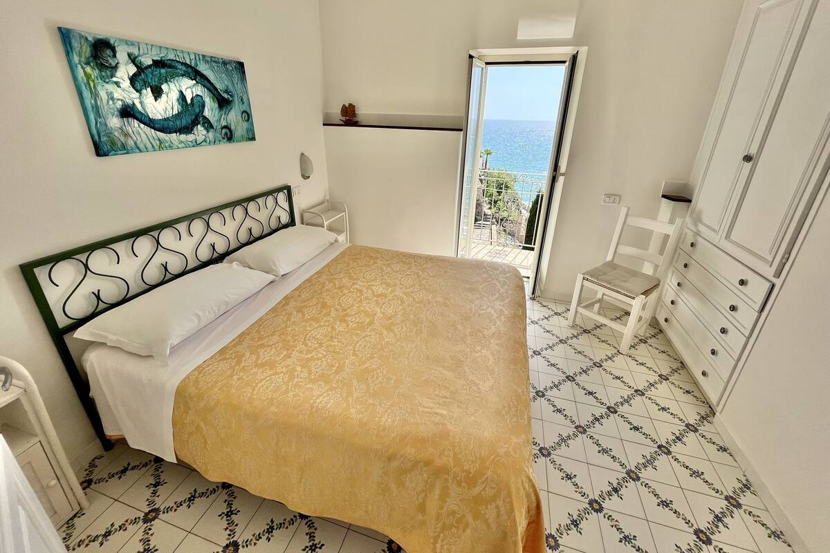 Ein Schlafzimmer mit einem Bett, einem Nachttisch, einer Kommode, einem Sessel, einem Gemälde und Meerblick durch eine geöffnete Tür.