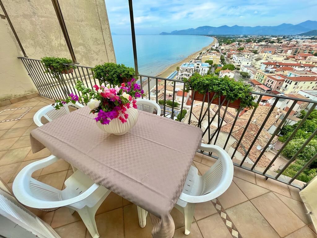 A balcony with a table and chairs, overlooking a coastal town and the sea.
