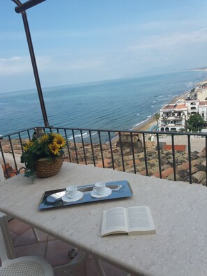 Un balcón con vista al mar, una mesa con una bandeja de tazas y platos, y un libro.