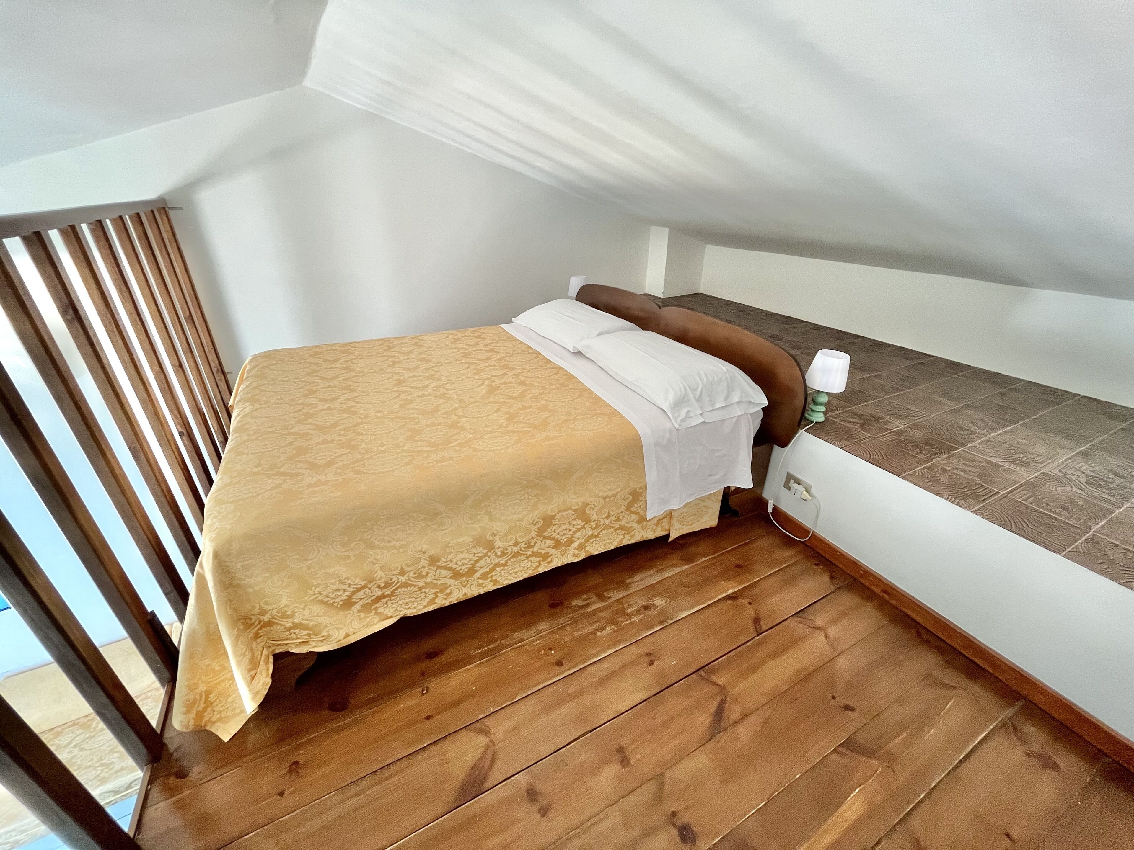 A small, single-bed room with a wooden floor, a white wall, and a window with blinds.
