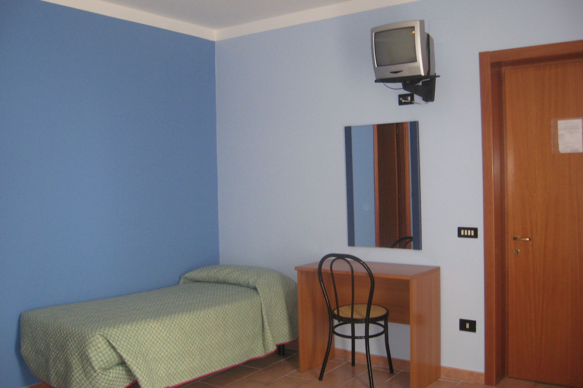 A small room with a bed, a desk, a chair, a television mounted on the wall, and a door.