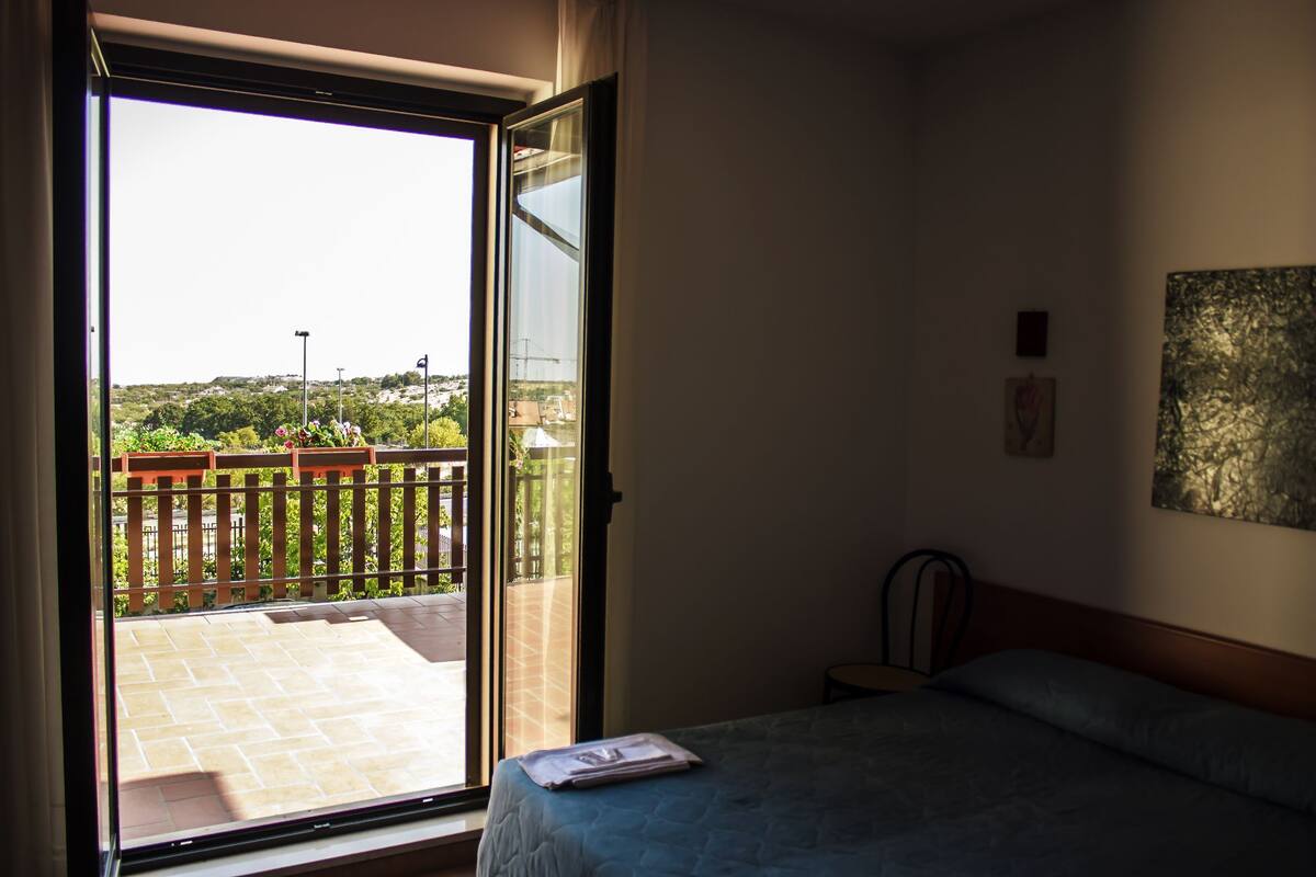 A hotel room with a bed, a view of a balcony with a wooden fence, and a painting on the wall.