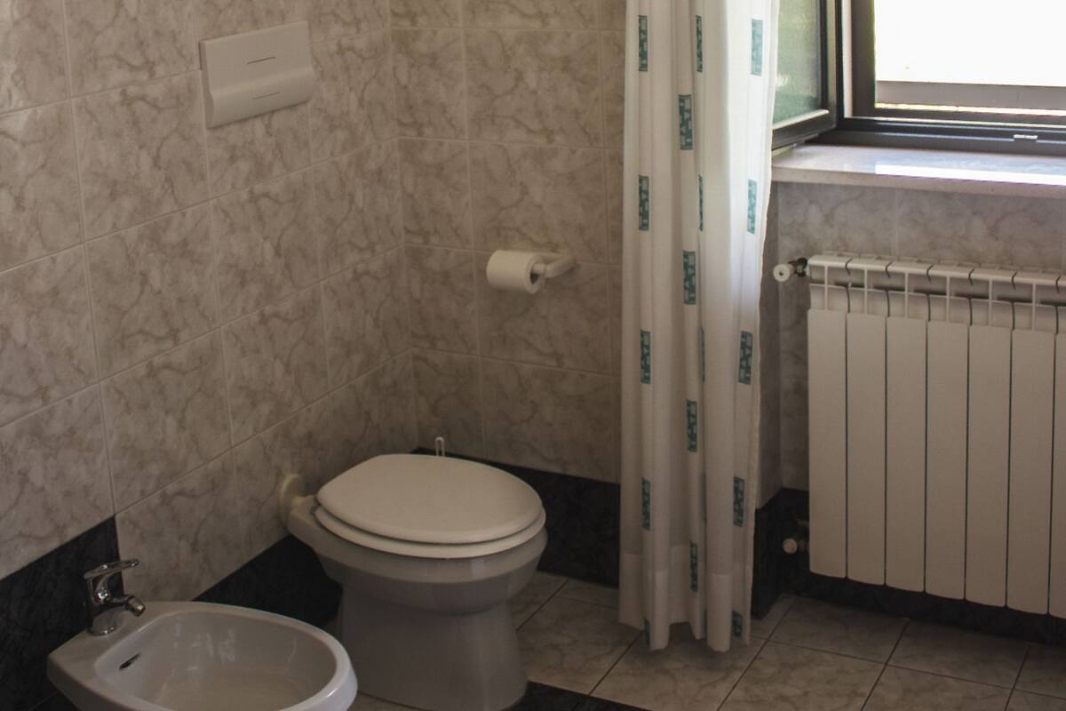 A bathroom with two toilets, a bidet, a radiator, and a window with curtains.