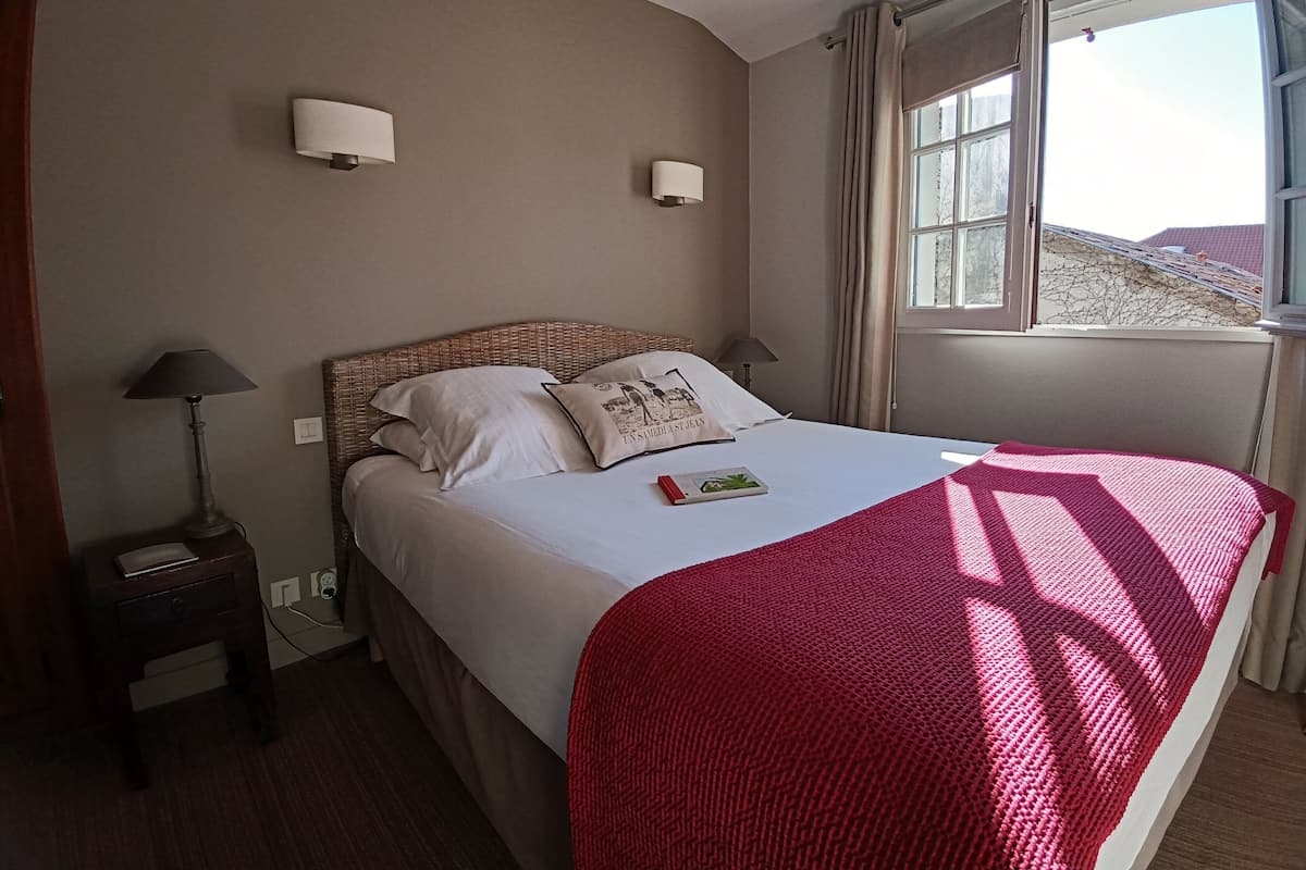 Een keurig opgemaakt bed met een rode deken, twee bedlampjes en een raam met gordijnen.