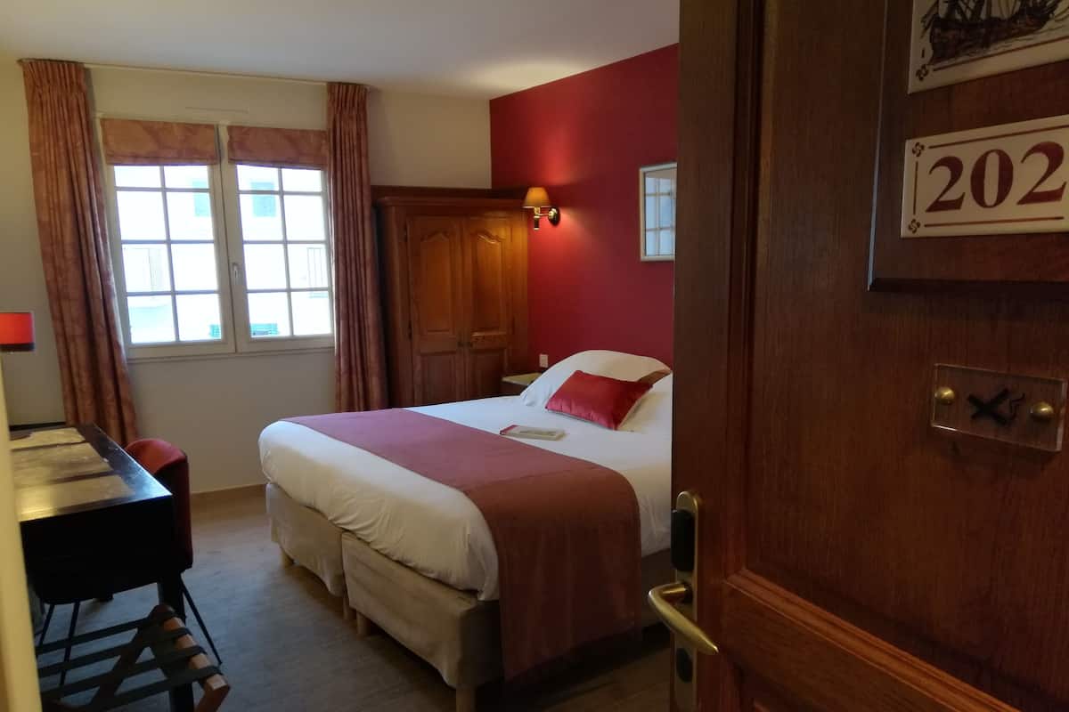 Een hotelkamer met een bed, een bureau en een stoel. Er is een raam met gordijnen, een deur met nummerbord, en een wandlamp.