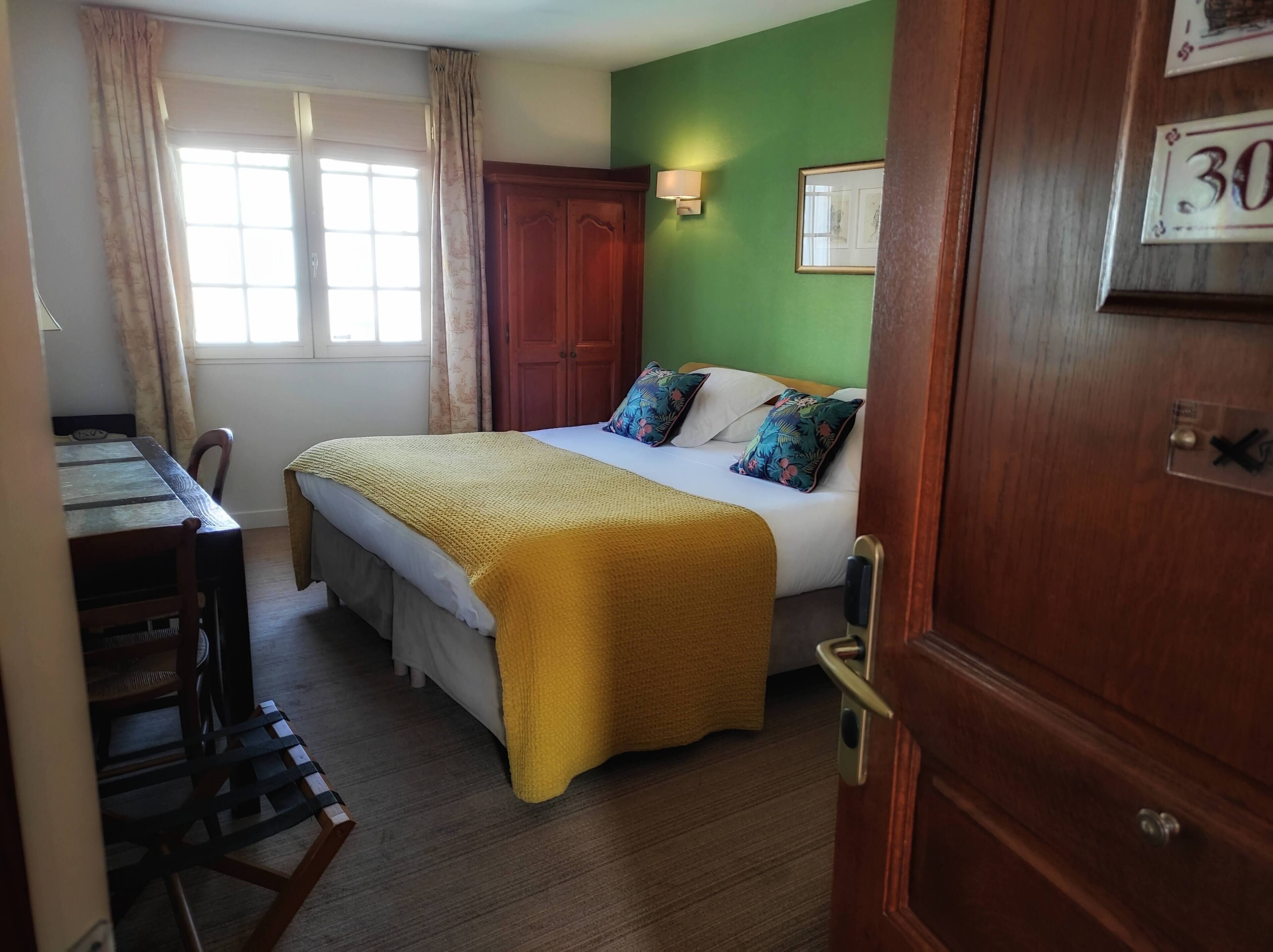 Ein Hotelzimmer mit Bett, Schreibtisch, Stuhl, einer Tür mit Nummer und einem Vorhangfenster.