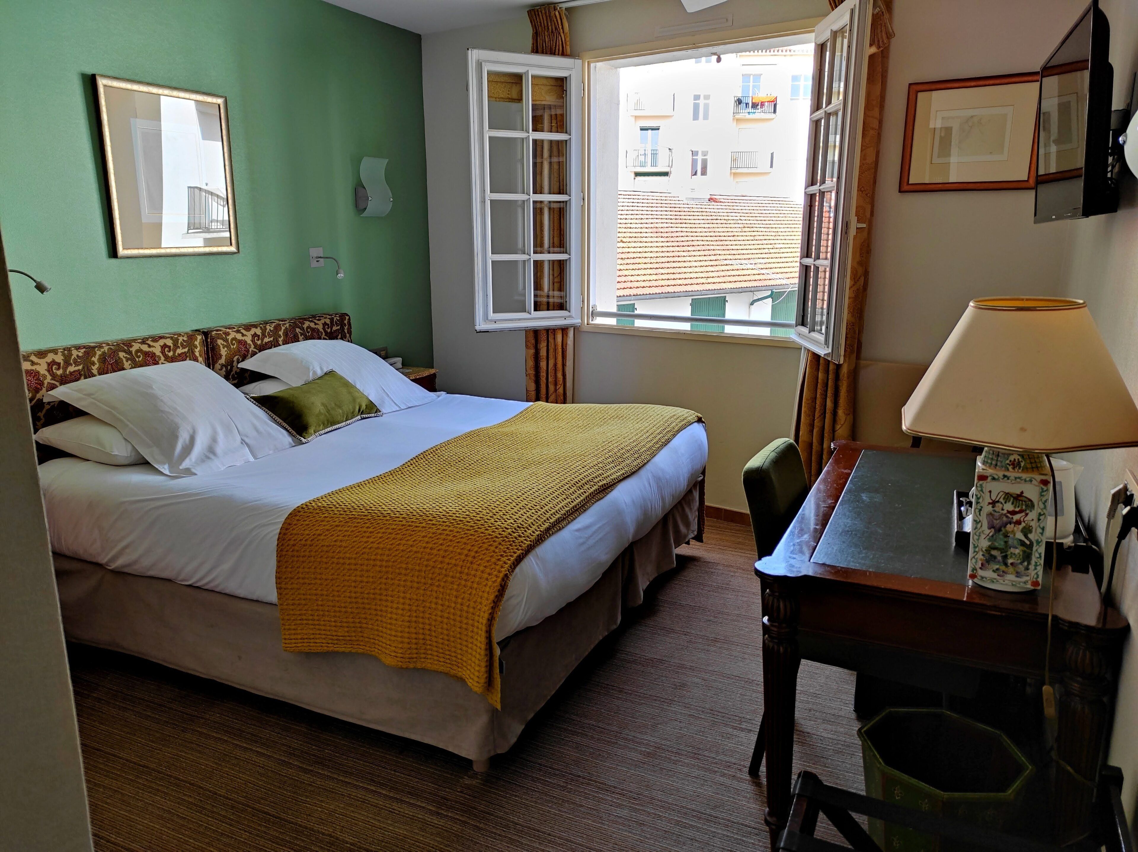 Ein Hotelzimmer mit Bett, Schreibtisch, Stuhl, einem Fenster mit Vorhängen und einer Wandlampe.