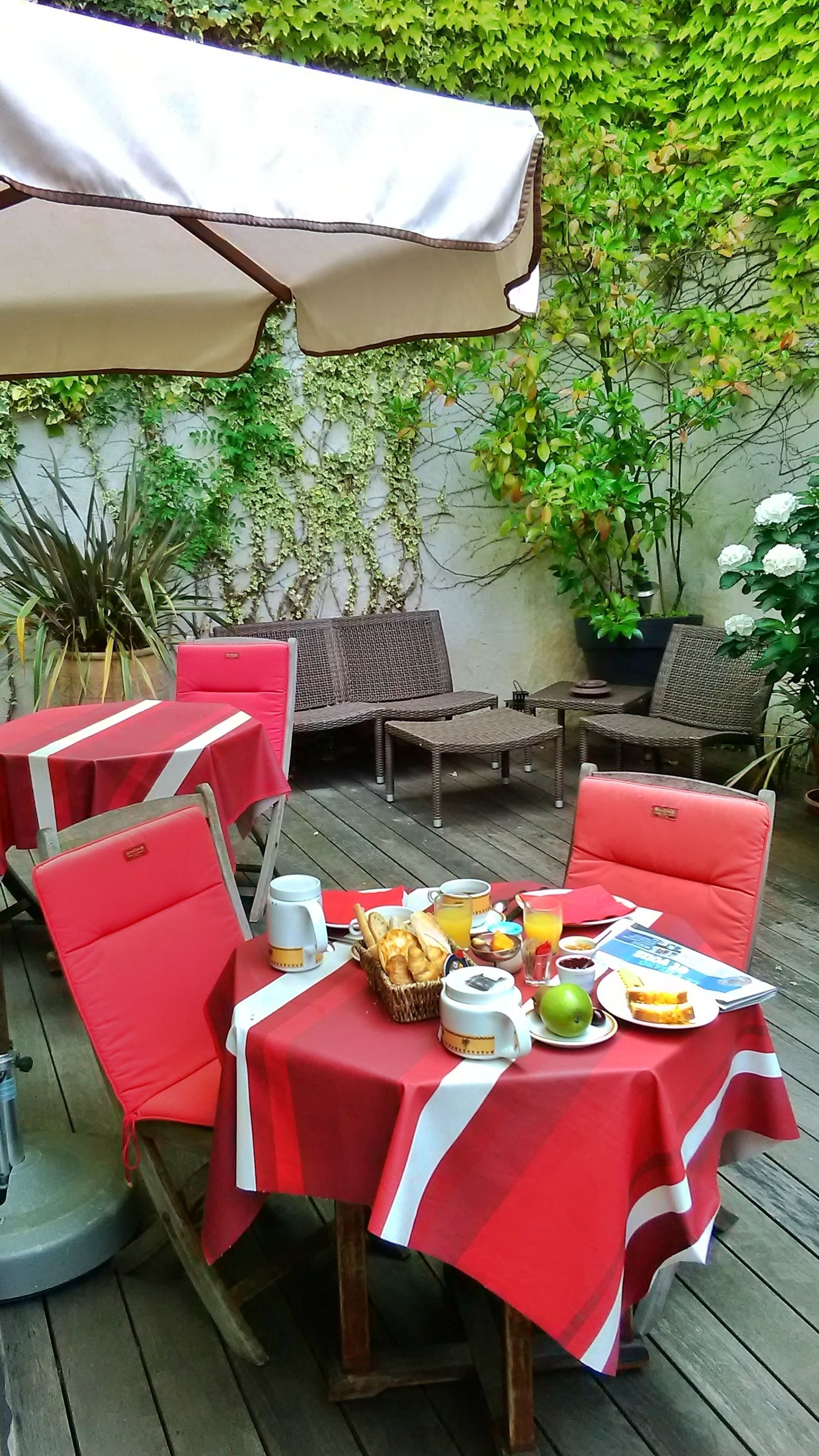 Un patio al aire libre con mesas y sillas cubiertas con manteles rojos y blancos, un sofá de mimbre y un paraguas blanco.
