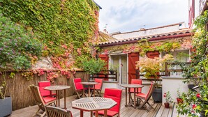 En udendørs terrasse med træborde og røde stole, omgivet af frodigt grønt og krukker med planter.