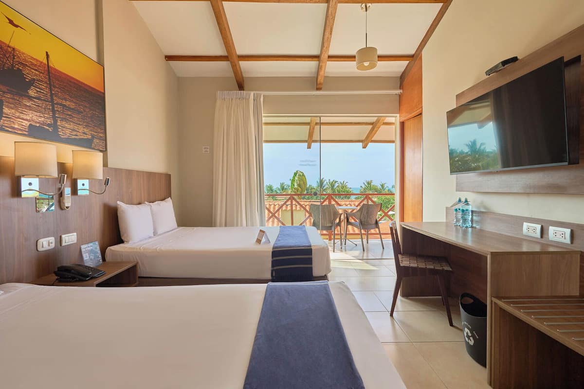 Quarto de hotel com duas camas, uma mesa de trabalho, televisão e varanda vista para o mar.