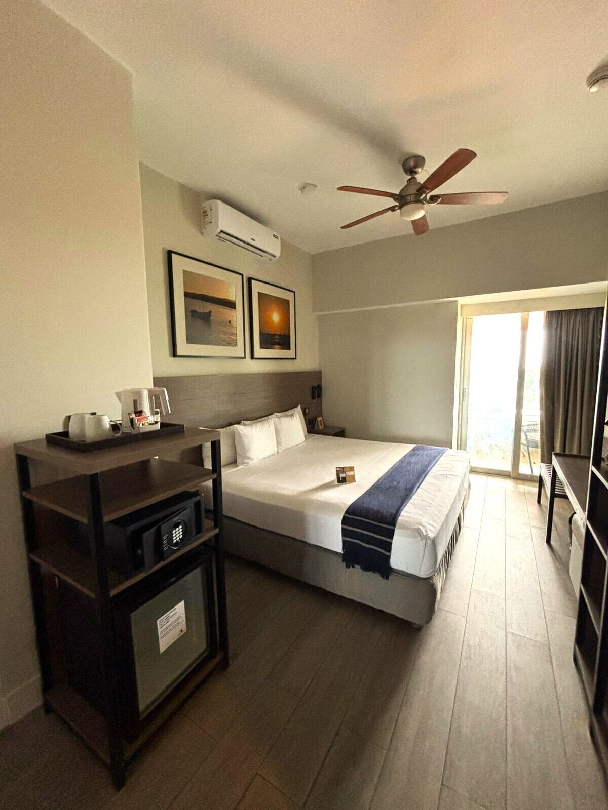 Habitación de hotel con cama, escritorio, ventilador de techo y ventana con cortinas.