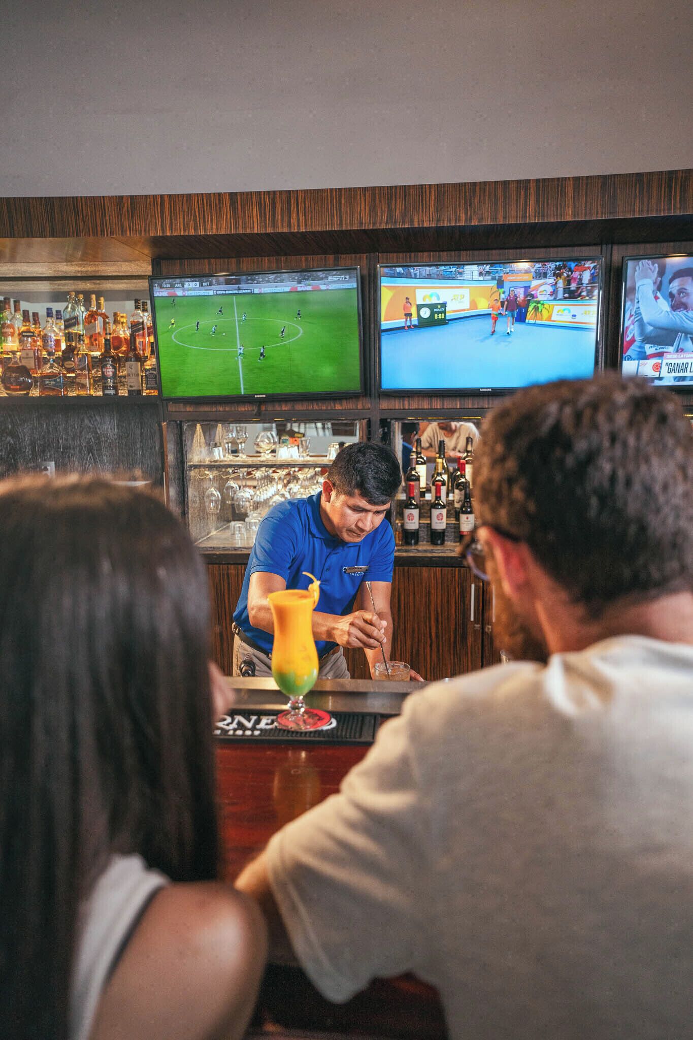 Eine Bar mit mehreren Bildschirmen, die Sportereignisse zeigen, einem Barkeeper, der einen Drink zubereitet, und einem Gast, der an der Bar sitzt.