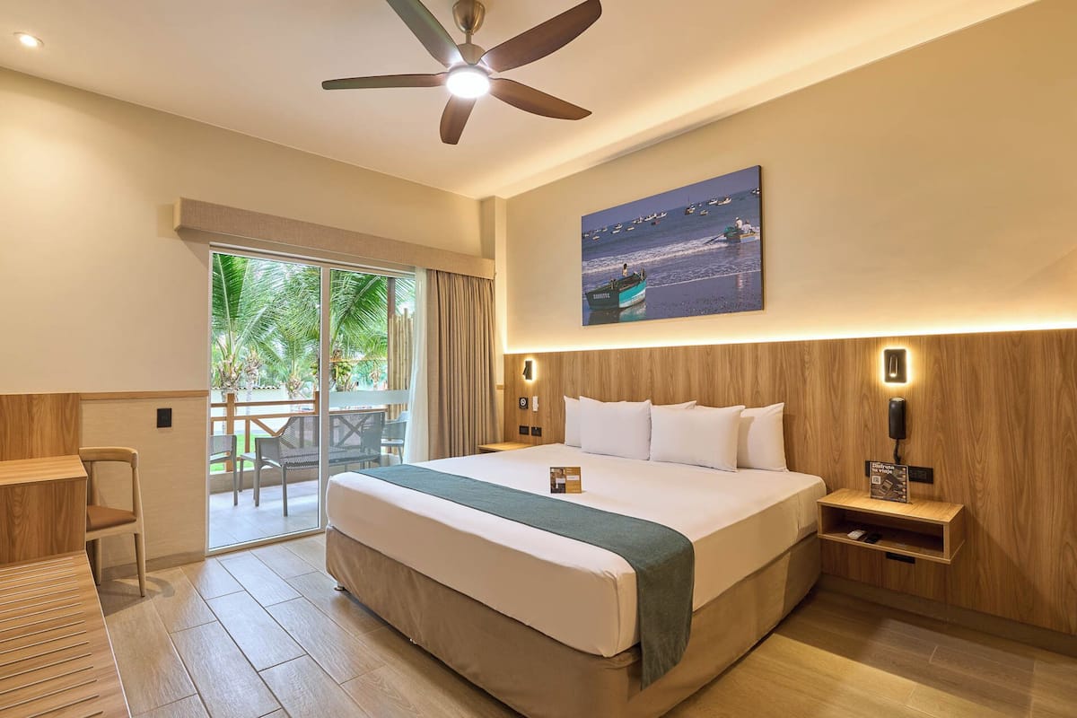 Quarto de hotel com cama, escrivaninha, cadeira, ventilador de teto, mesa de cabeceira, telefone e pintura de barcos na parede.