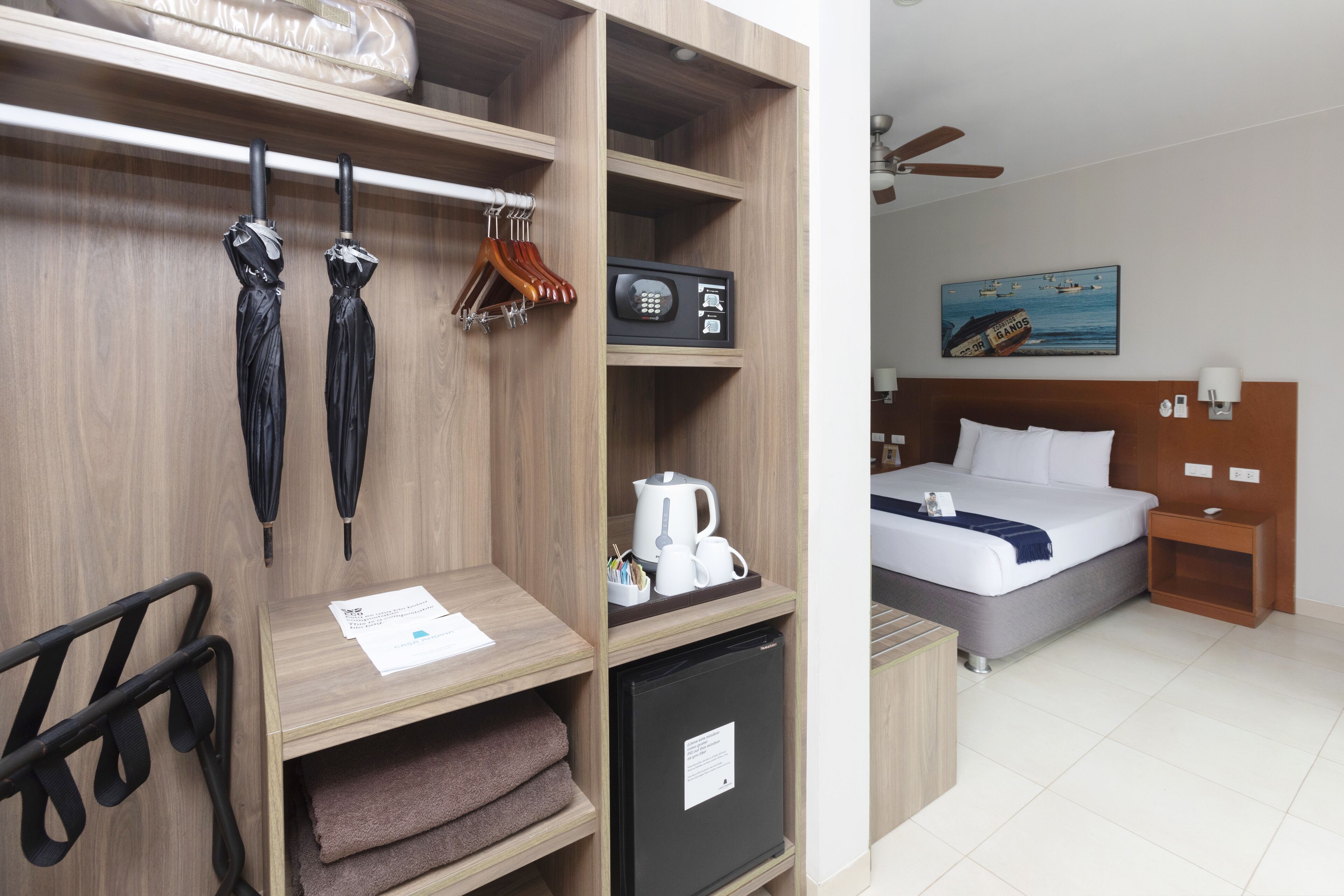 A hotel room with a bed, a nightstand, a desk with a kettle, and a wardrobe with hangers.