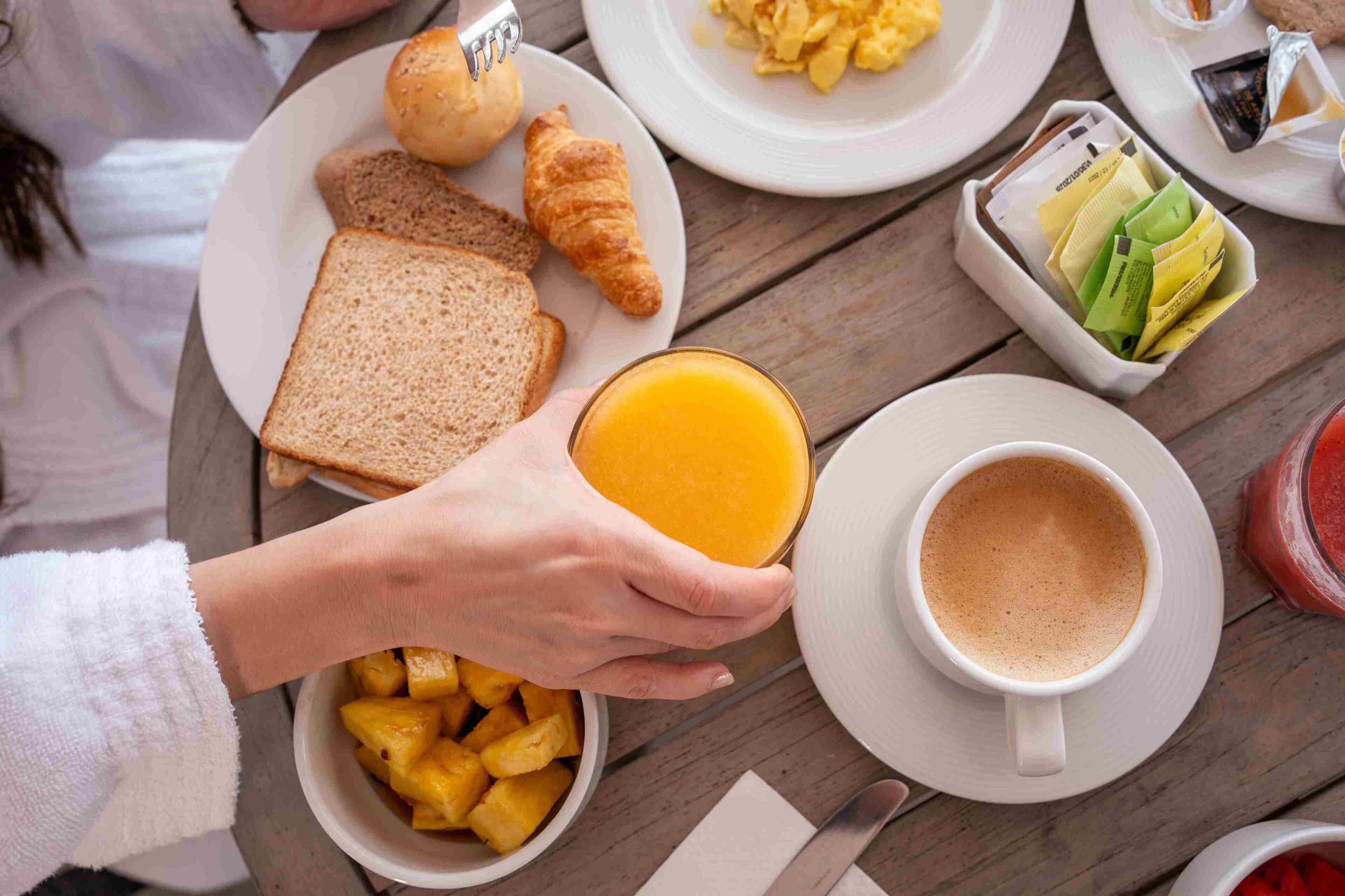 Mesa de madeira com pão tostado, croissants, frutas e uma xícara de café.