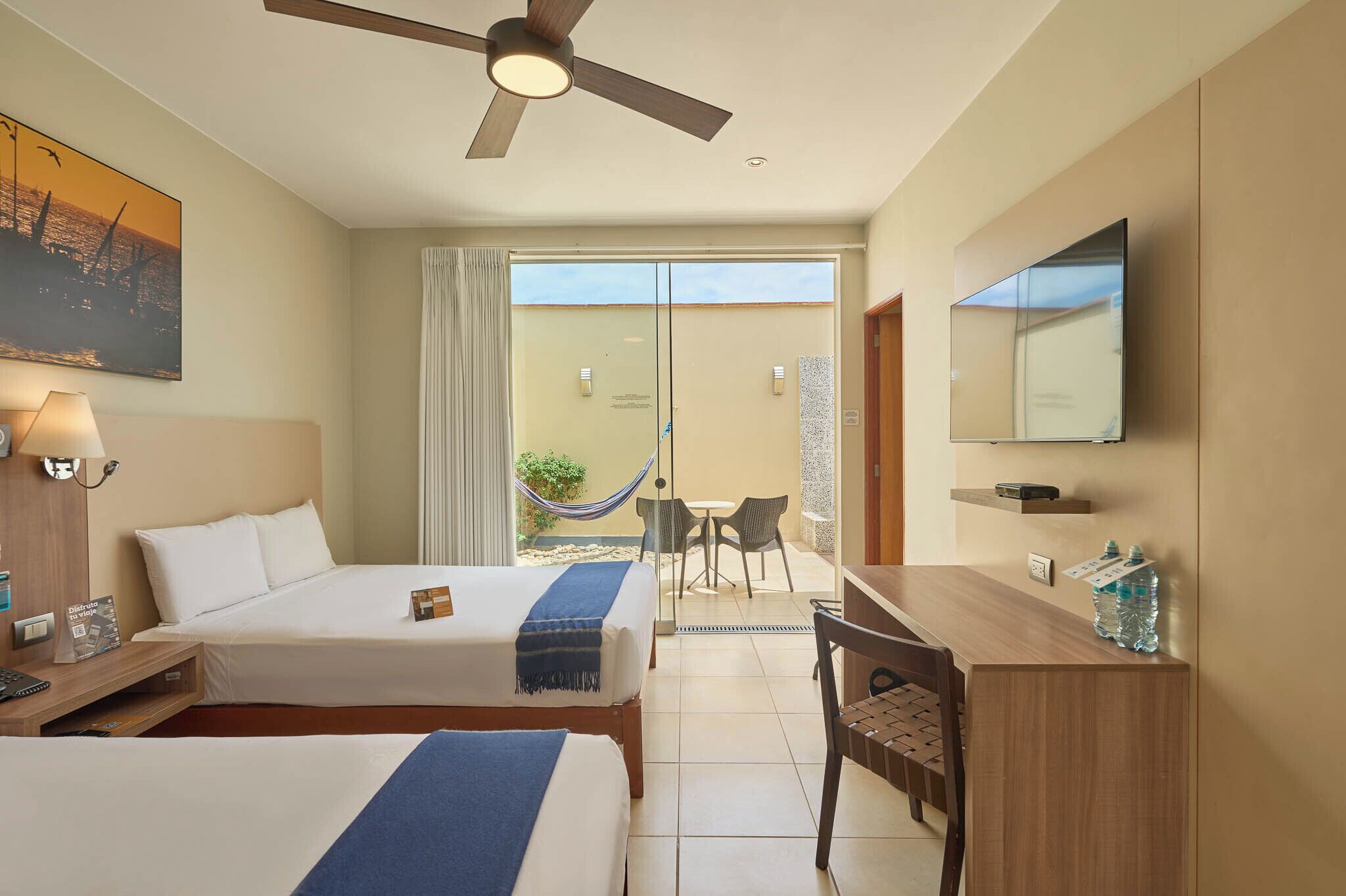 Habitación de hotel con dos camas, un escritorio, una silla, un televisor, un ventilador de techo y un balcón con una hamaca.