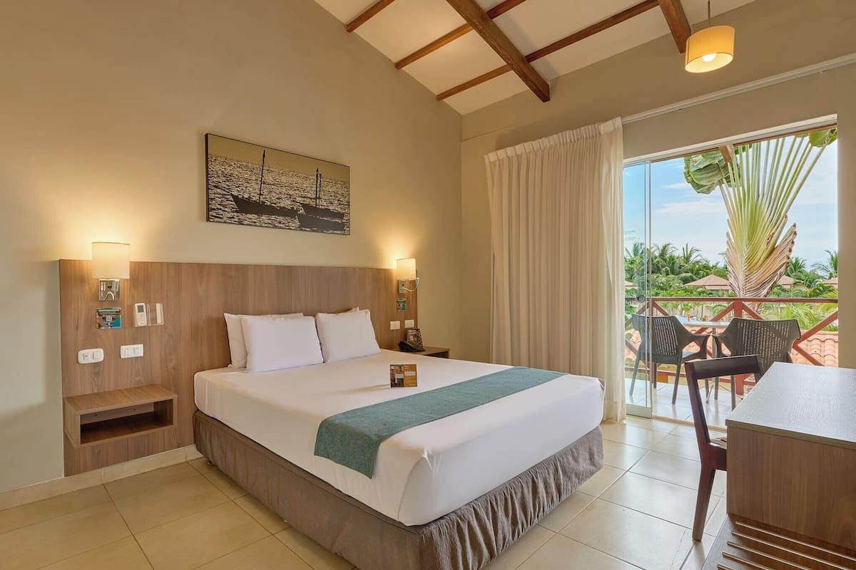 Quarto de hotel com uma cama grande, cabeceira de madeira, um quadro, mesa de cabeceira com abajur, escrivaninha com cadeiras e varanda com vista para palmeiras.