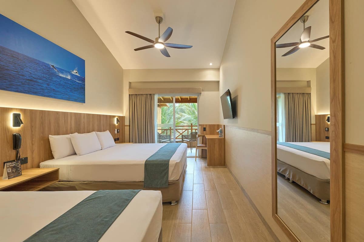 Quarto de hotel com duas camas, um ventilador de teto, televisão e uma janela grande com vista para o mar.