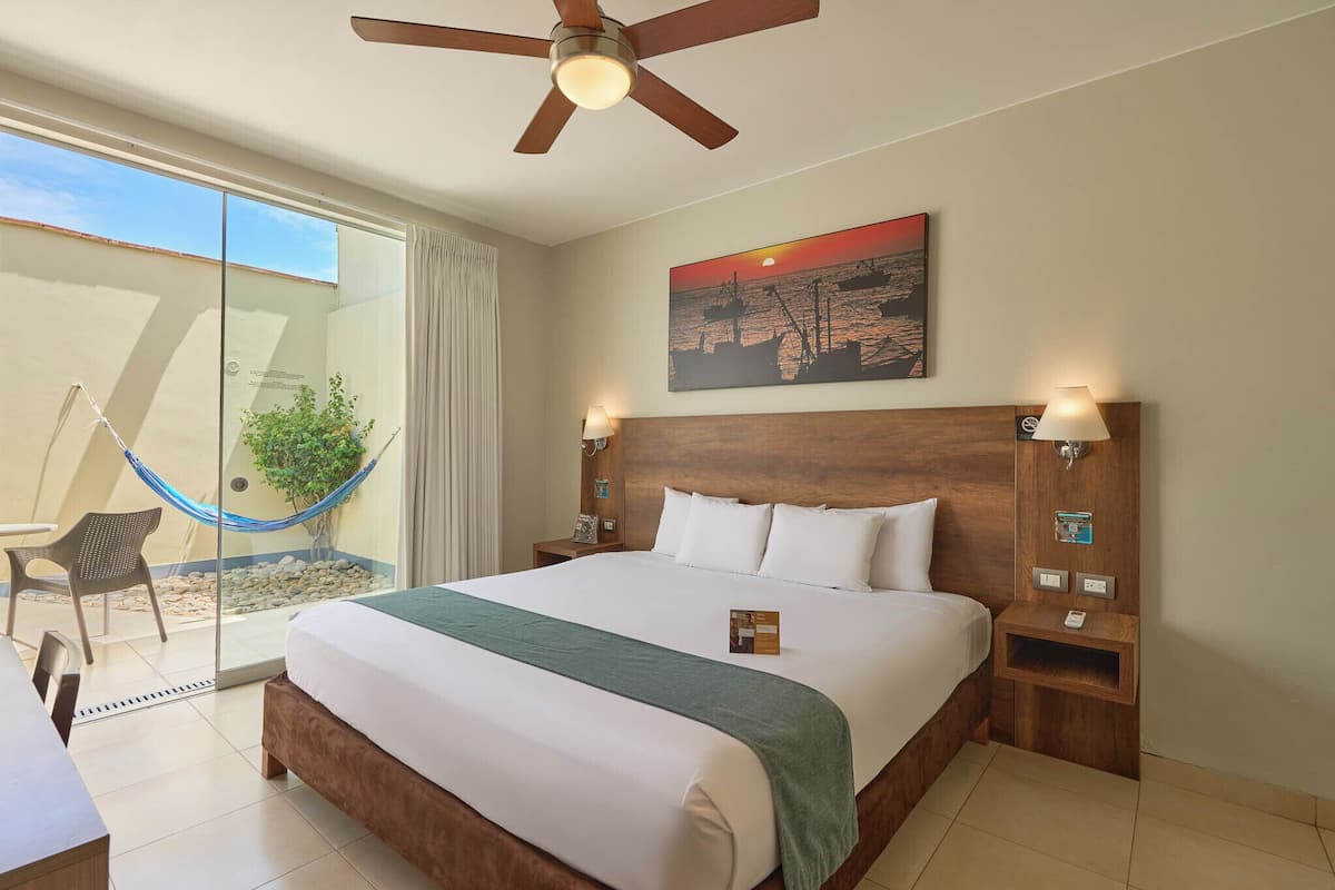 Quarto de hotel com uma cama grande, um ventilador de teto, uma rede, uma porta de correr de vidro e uma pintura de um pôr do sol.