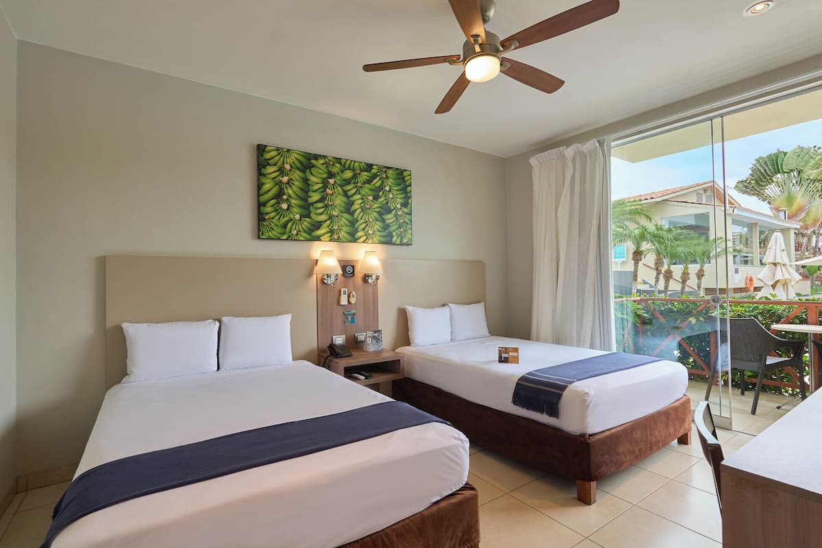 Quarto de hotel com duas camas, um ventilador de teto, uma pintura de bananas e vista para o exterior.
