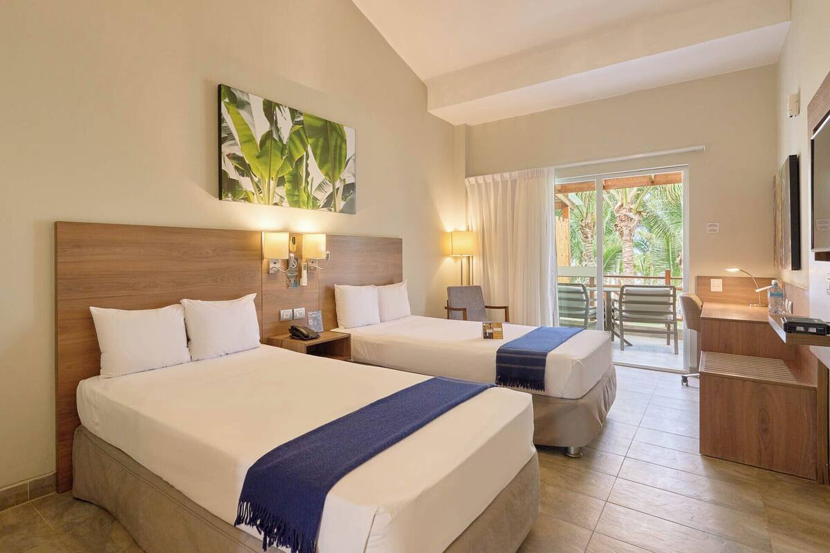 Quarto de hotel com duas camas, varanda com vista para palmeiras, secretária com cadeira e um quadro na parede.
