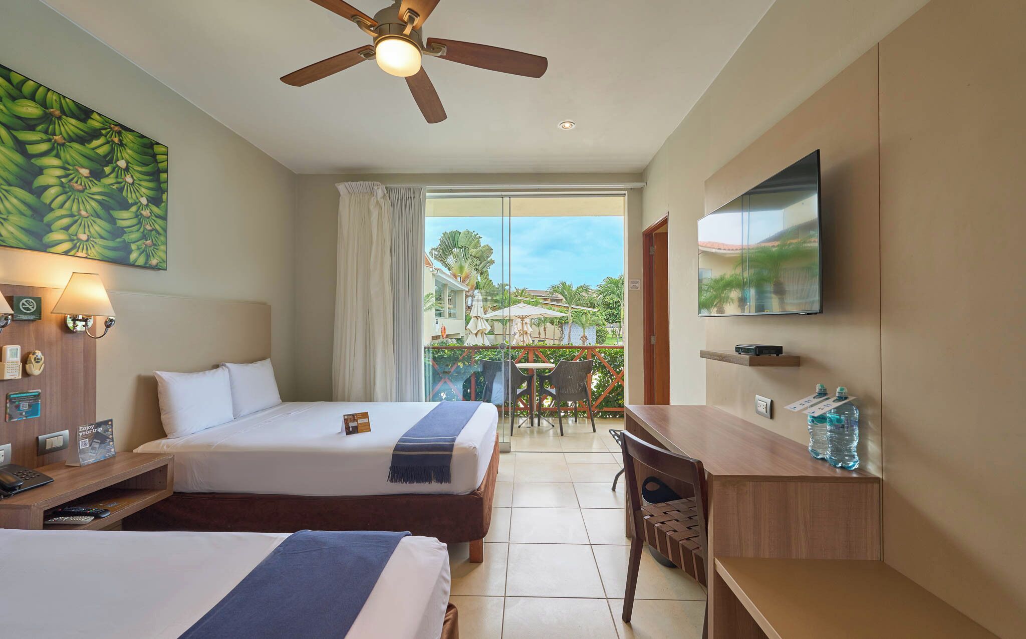 Habitación de hotel con dos camas, un escritorio, una silla, ventilador de techo, televisor y balcón con mobiliario exterior.
