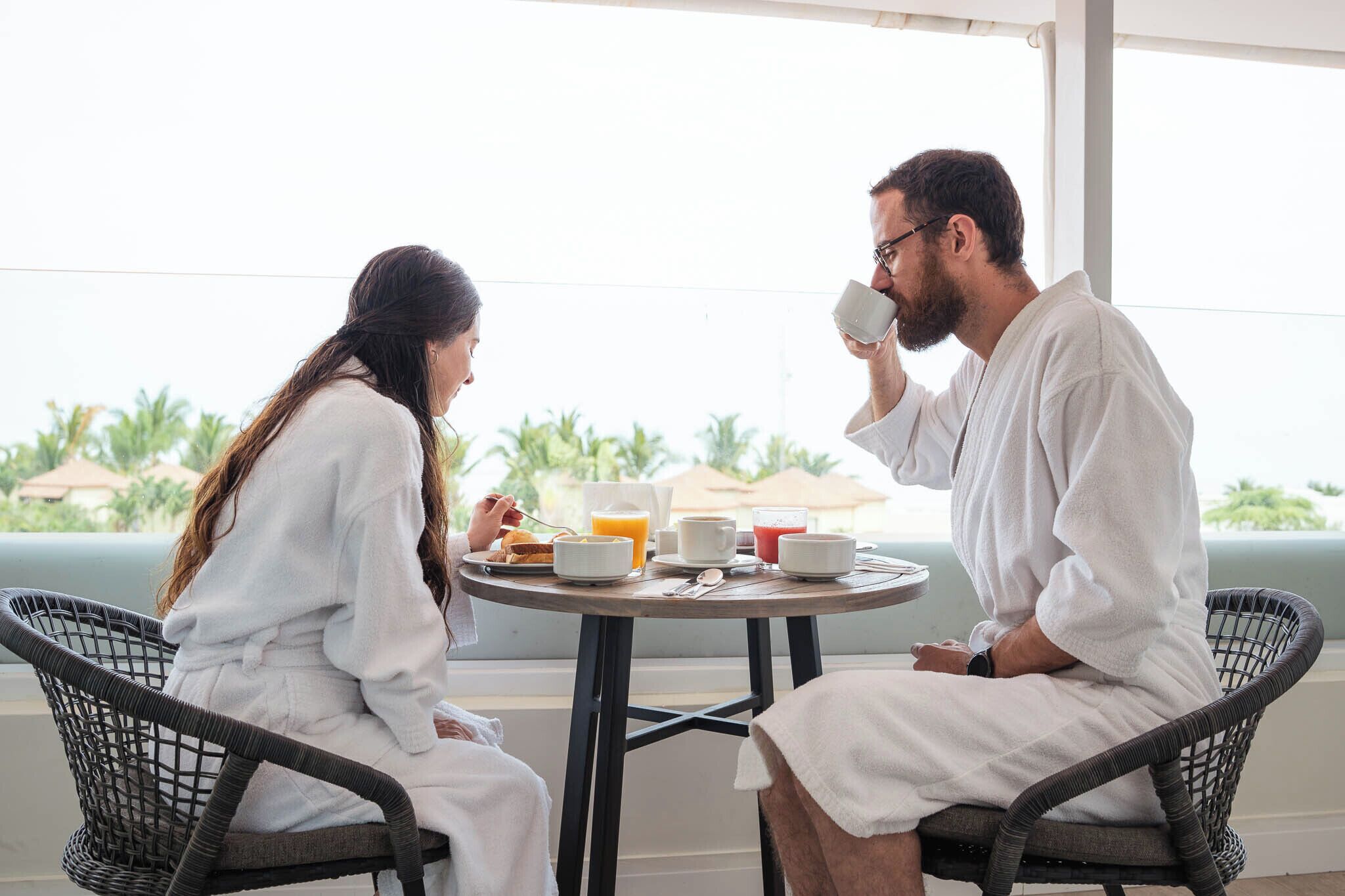 Deux personnes vêtues de robes blanches sont assises autour d’une table ronde, devant un petit-déjeuner, dans une pièce d’où l’on peut admirer des palmiers.