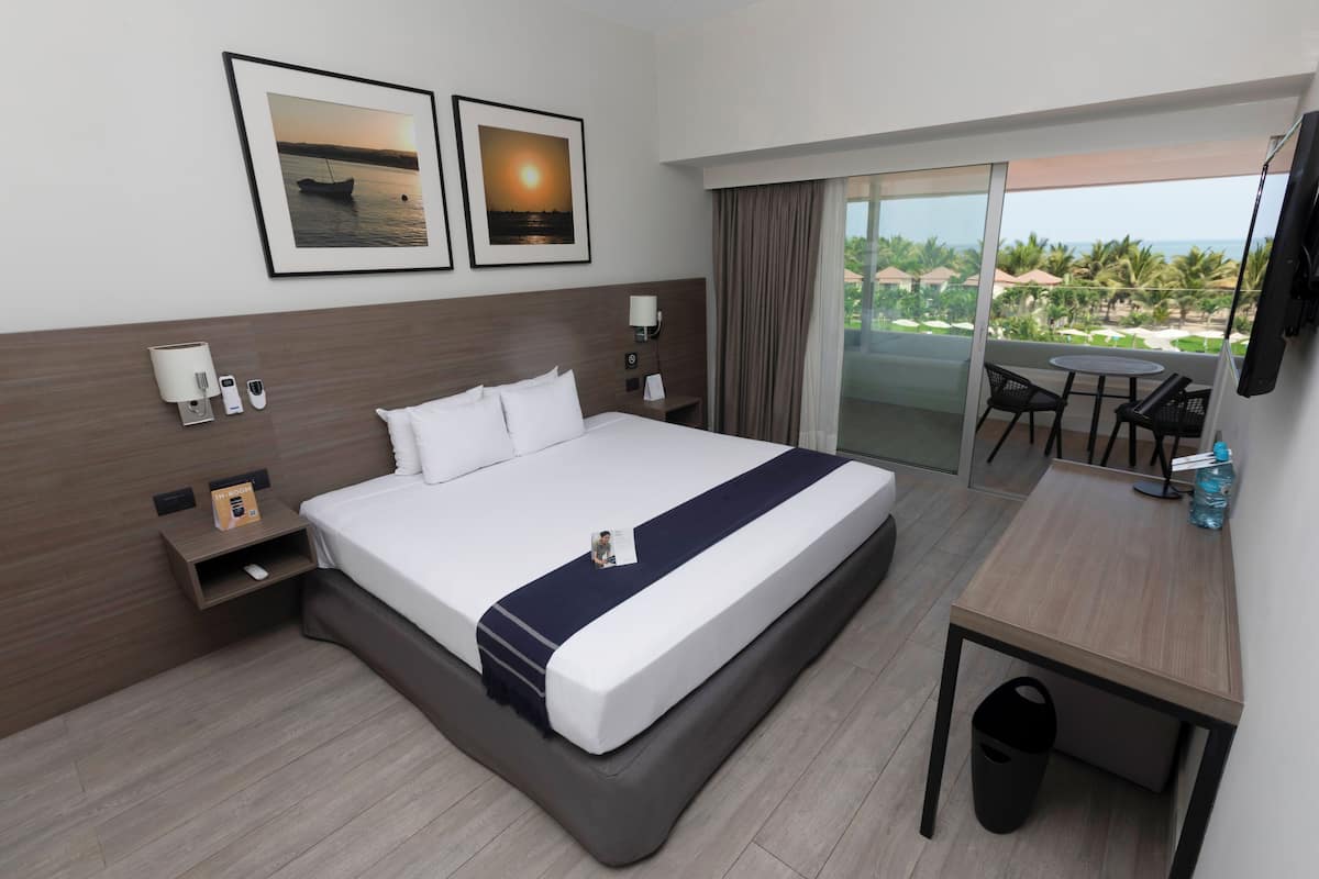 Quarto de hotel com uma cama grande, uma mesa de trabalho, uma cadeira, televisão e varanda com vista para o mar.